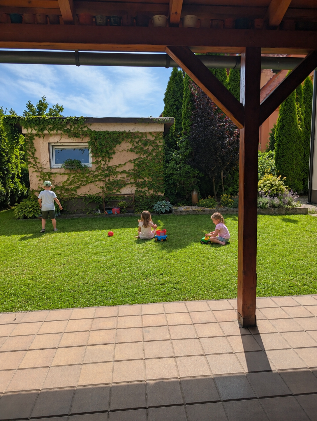 Děti si hrají na zahradě pod dřevěnou pergolou, na trávníku s hračkami.
